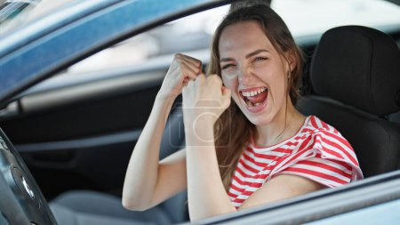 Téléchargez les photos : Jeune femme blonde souriant confiant assis sur la voiture avec l'expression gagnante à la rue - en image libre de droit