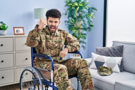 Foto de Hombre árabe vistiendo uniforme camuflaje del ejército sentado en silla de ruedas enojado y loco levantando el puño frustrado y furioso mientras gritaba con ira. rabia y concepto agresivo. - Imagen libre de derechos