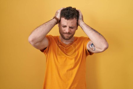 Foto de Young hispanic man standing over yellow background suffering from headache desperate and stressed because pain and migraine. hands on head. - Imagen libre de derechos