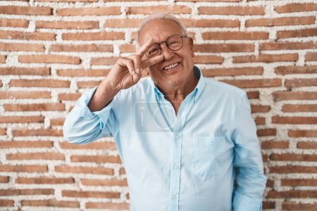 Foto de Hombre mayor con el pelo gris de pie sobre la pared de ladrillos haciendo símbolo de paz con los dedos sobre la cara, sonriendo alegre mostrando la victoria - Imagen libre de derechos