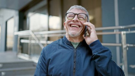 Foto de Hombre de pelo gris de mediana edad sonriendo confiado hablando en el teléfono inteligente en la calle - Imagen libre de derechos