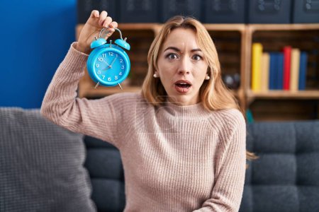 Photo for Hispanic woman holding alarm clock scared and amazed with open mouth for surprise, disbelief face - Royalty Free Image