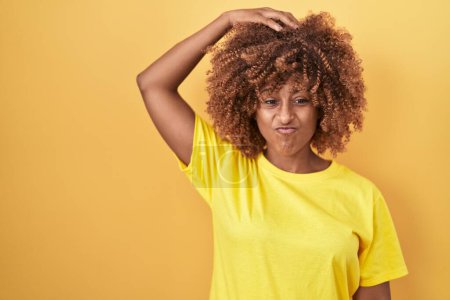 Photo for Young hispanic woman with curly hair standing over yellow background confuse and wonder about question. uncertain with doubt, thinking with hand on head. pensive concept. - Royalty Free Image