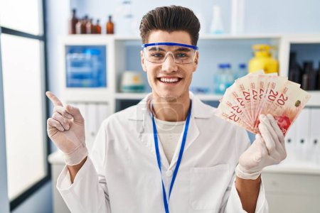 Photo for Young hispanic man working at scientist laboratory holding shekels smiling happy pointing with hand and finger to the side - Royalty Free Image