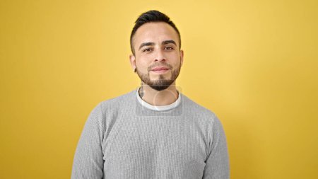 Hispanic man standing with serious expression over isolated yellow background