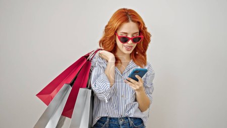 Photo for Young redhead woman using smartphone holding shopping bags over isolated white background - Royalty Free Image