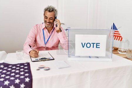 Téléchargez les photos : Moyen-âge homme aux cheveux gris président de table électorale écrivant sur presse-papiers parler sur smartphone au collège électoral - en image libre de droit