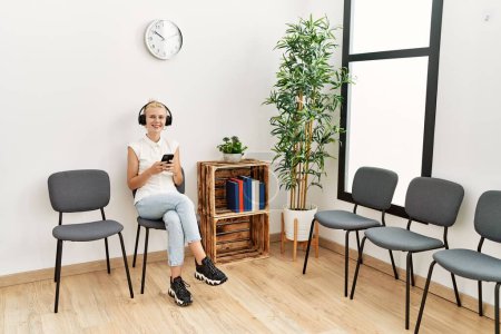 Foto de Mujer rubia joven usando teléfono inteligente y auriculares sentados en la silla en la sala de espera - Imagen libre de derechos
