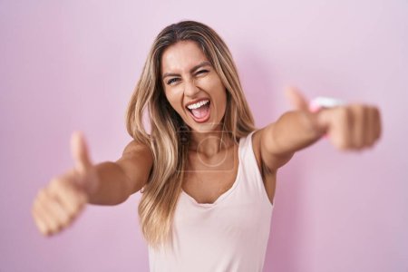 Photo for Young blonde woman standing over pink background approving doing positive gesture with hand, thumbs up smiling and happy for success. winner gesture. - Royalty Free Image