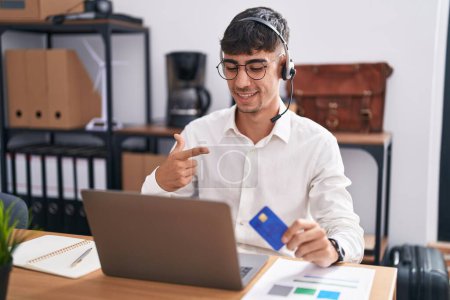 Téléchargez les photos : Jeune homme hispanique travaillant à l'aide d'un ordinateur portable tenant la carte de crédit gai avec un sourire sur le visage pointant avec la main et le doigt vers le côté avec une expression heureuse et naturelle - en image libre de droit
