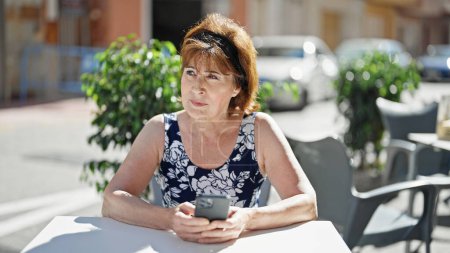 Téléchargez les photos : Femme d'âge moyen utilisant smartphone assis sur la table à la terrasse du café - en image libre de droit