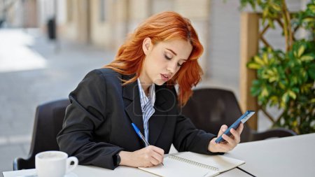 Téléchargez les photos : Jeune rousse femme d'affaires utilisant un smartphone prenant des notes à la terrasse du café - en image libre de droit