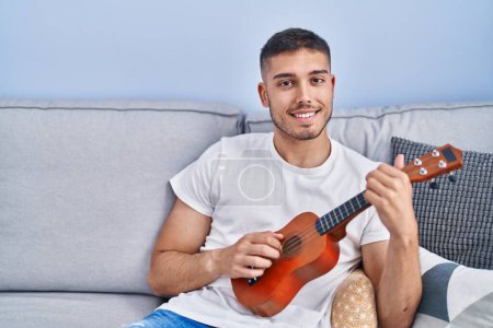Foto de Young hispanic man playing ukelele sitting on sofa at home - Imagen libre de derechos