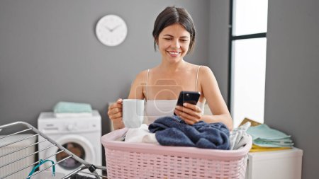 Photo for Young beautiful hispanic woman using smartphone drinking coffee leaning on clothesline at laundry room - Royalty Free Image
