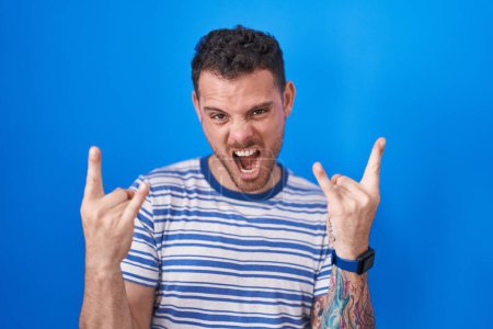 Photo for Young hispanic man standing over blue background shouting with crazy expression doing rock symbol with hands up. music star. heavy music concept. - Royalty Free Image