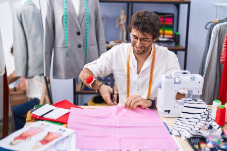 Téléchargez les photos : Jeune homme hispanique tailleur sourire tissu de coupe confiant à l'usine de vêtements - en image libre de droit