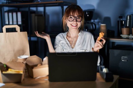 Photo for Young beautiful woman working using computer laptop and eating delivery food smiling cheerful presenting and pointing with palm of hand looking at the camera. - Royalty Free Image