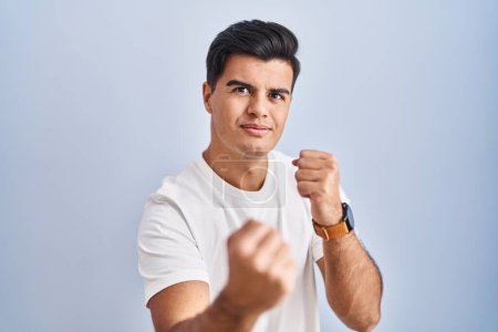Photo for Hispanic man standing over blue background ready to fight with fist defense gesture, angry and upset face, afraid of problem - Royalty Free Image