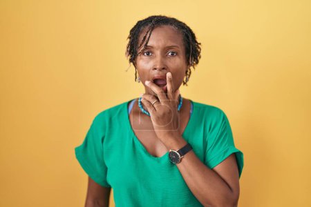 Foto de Mujer africana con rastas de pie sobre fondo amarillo que se ve fascinada con la incredulidad, sorpresa y expresión asombrada con las manos en la barbilla - Imagen libre de derechos