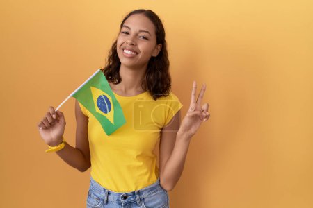Foto de Mujer hispana joven sosteniendo bandera brasileña sonriendo mirando a la cámara mostrando los dedos haciendo señal de victoria. número dos. - Imagen libre de derechos