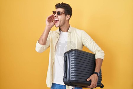 Foto de Joven hispano sosteniendo la maleta yendo de vacaciones de verano gritando y gritando fuerte a un lado con la mano en la boca. concepto de comunicación. - Imagen libre de derechos