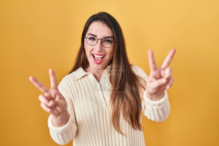 Foto de Mujer hispana joven de pie sobre fondo amarillo sonriendo mirando a la cámara mostrando los dedos haciendo señal de victoria. número dos. - Imagen libre de derechos