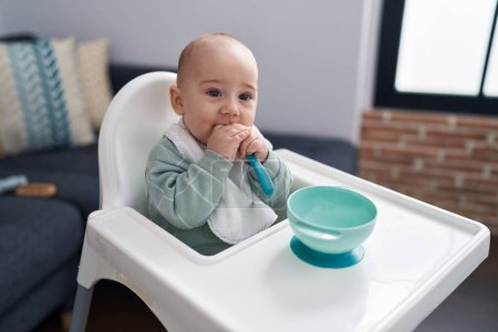 Foto de Adorable bebé caucásico sentado en silla alta chupando cuchara en casa - Imagen libre de derechos
