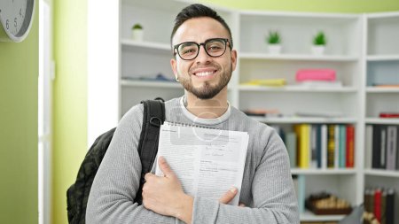Téléchargez les photos : Hispanique homme étudiant souriant confiant tenant devoirs à la bibliothèque universitaire - en image libre de droit