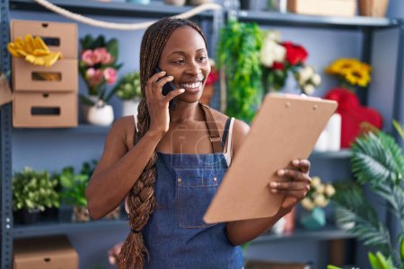 Téléchargez les photos : Afro-américaine femme fleuriste parler sur smartphone lecture presse-papiers à la boutique de fleurs - en image libre de droit