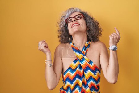 Téléchargez les photos : Femme d'âge moyen avec les cheveux gris debout sur fond jaune célébrant surpris et étonnés pour le succès avec les bras levés et les yeux fermés. concept gagnant. - en image libre de droit