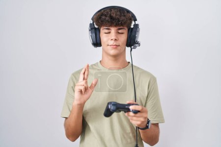 Téléchargez les photos : Adolescent hispanique jouant jeu vidéo tenant contrôleur geste doigt croisé sourire avec espoir et les yeux fermés. chance et concept superstitieux. - en image libre de droit