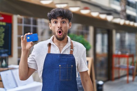 Photo for Arab man with beard wearing waiter apron at restaurant terrace holding credit card scared and amazed with open mouth for surprise, disbelief face - Royalty Free Image