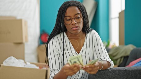 Photo for African american woman sitting on sofa counting dollars looking upset at new home - Royalty Free Image