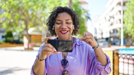 Foto de Joven hermosa mujer latina viendo video en smartphone con gesto ganador en el parque - Imagen libre de derechos