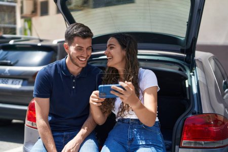 Téléchargez les photos : Jeune couple hispanique assis sur le coffre de la voiture regarder la vidéo sur smartphone à la rue - en image libre de droit