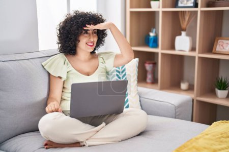 Photo for Young brunette woman with curly hair using laptop sitting on the sofa at home very happy and smiling looking far away with hand over head. searching concept. - Royalty Free Image