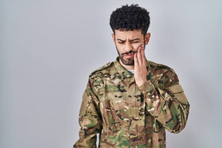 Téléchargez les photos : Homme arabe portant un uniforme de camouflage militaire touchant la bouche avec la main avec une expression douloureuse à cause de maux de dents ou de maladies dentaires sur les dents. dentiste - en image libre de droit