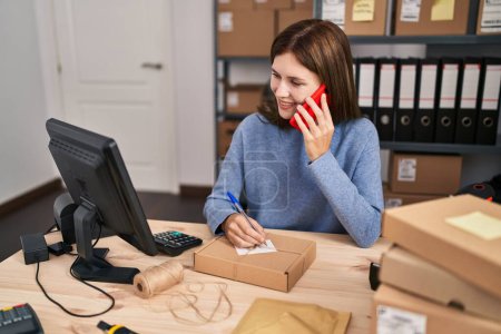 Foto de Mujer rubia joven comercio electrónico trabajador de negocios hablando en la escritura de teléfonos inteligentes en el paquete en la oficina - Imagen libre de derechos