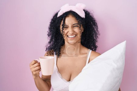 Foto de Mujer hispana con el pelo rizado usando pijama bebiendo una taza de café sonriendo y riendo en voz alta porque divertida broma loca. - Imagen libre de derechos