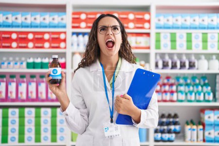 Photo for Young hispanic woman working at pharmacy drugstore holding syrup angry and mad screaming frustrated and furious, shouting with anger looking up. - Royalty Free Image