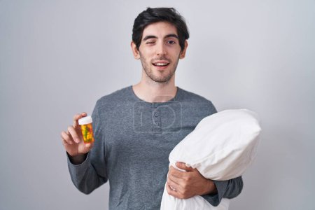 Téléchargez les photos : Jeune homme hispanique portant pyjama étreignant oreiller tenant pilules clin d'oeil en regardant la caméra avec expression sexy, visage joyeux et heureux. - en image libre de droit
