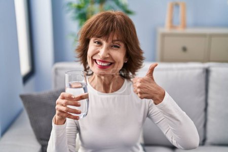 Foto de Mujer de mediana edad beber vaso de agua sonriendo feliz y positivo, pulgar hacia arriba haciendo excelente y signo de aprobación - Imagen libre de derechos