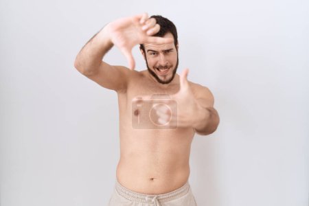 Téléchargez les photos : Jeune homme hispanique debout torse nu sur fond blanc souriant faisant cadre avec les mains et les doigts avec le visage heureux. concept de créativité et photographie. - en image libre de droit