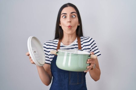 Photo for Young brunette woman wearing apron holding cooking pot making fish face with mouth and squinting eyes, crazy and comical. - Royalty Free Image