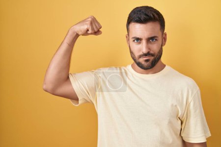 Foto de Hombre hispano guapo de pie sobre fondo amarillo fuerte persona mostrando el músculo del brazo, confiado y orgulloso de poder - Imagen libre de derechos