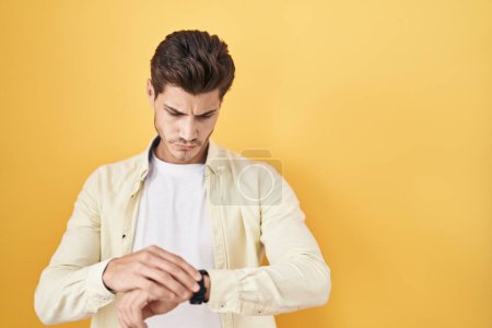 Téléchargez les photos : Jeune homme hispanique debout sur fond jaune vérifiant l'heure sur la montre-bracelet, détendu et confiant - en image libre de droit