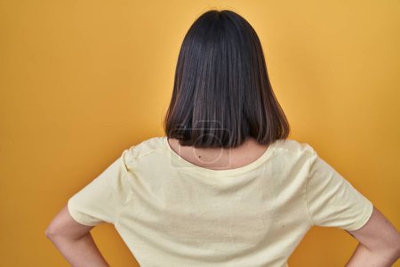 Foto de Chica hispana con camiseta casual sobre fondo amarillo de pie hacia atrás mirando hacia otro lado con los brazos en el cuerpo - Imagen libre de derechos