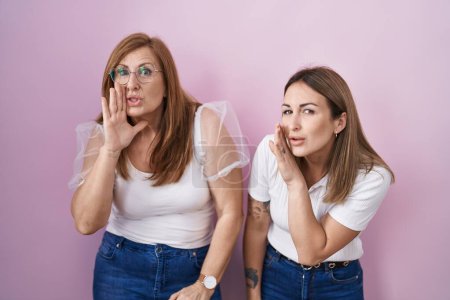 Foto de Madre e hija hispanas vistiendo camiseta blanca casual sobre fondo rosa mano en boca contando rumores secretos, susurrando conversación maliciosa - Imagen libre de derechos