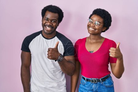 Photo for Young african american couple standing over pink background doing happy thumbs up gesture with hand. approving expression looking at the camera showing success. - Royalty Free Image
