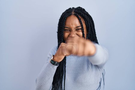 Téléchargez les photos : Femme afro-américaine debout sur fond bleu coup de poing poing pour se battre, attaque agressive et en colère, menace et violence - en image libre de droit